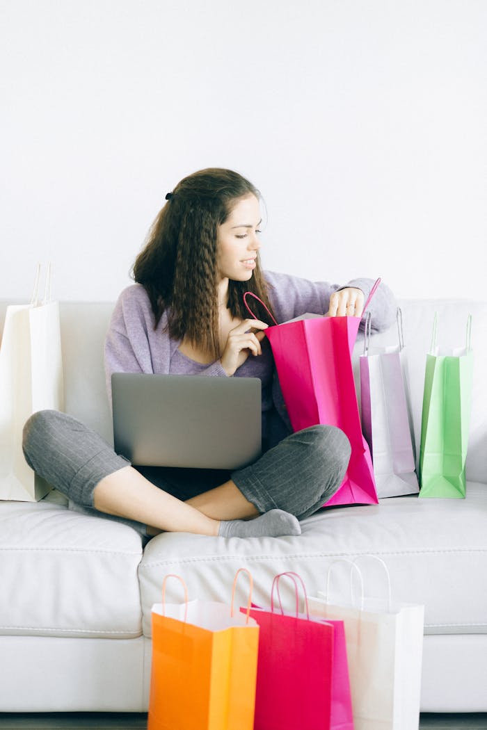 Woman on couch enjoying online shopping surrounded by colorful shopping bags and laptop.